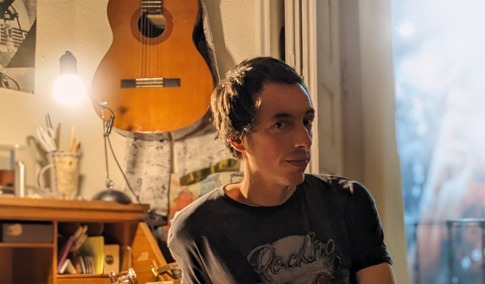 Alberto en su estudio con su guitarra colgada en el fondo, mirando a la cámera, con algo que puede ser una sonrisa
